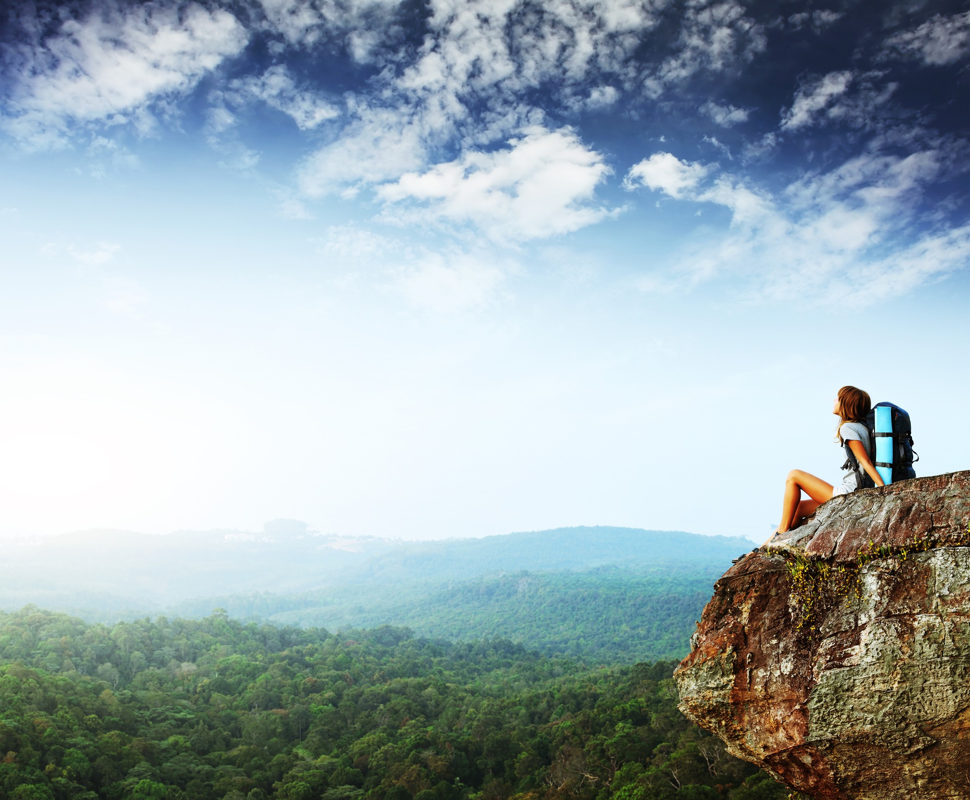 Young woman with backpack sitting on cliff and looking to a sky